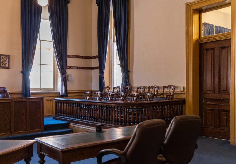 inside of an empty jury room with many wooden chairs in front of the jury box, an empty desk with two chairs in the foreground, and a witness stand.